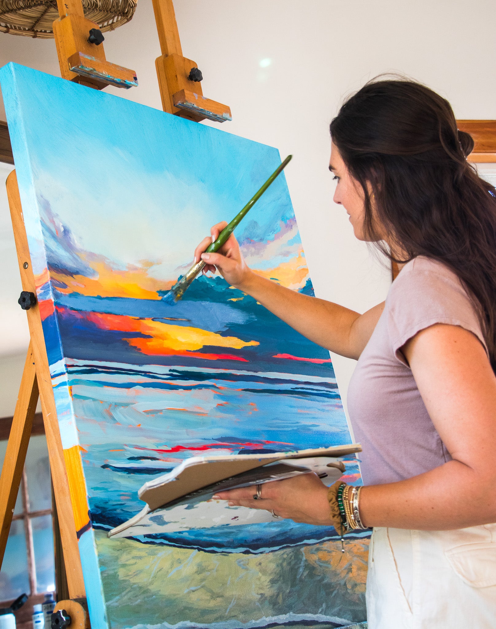 A woman painting a sunset seascape on a canvas set on an easel, holding a paintbrush and palette in a bright indoor studio.
